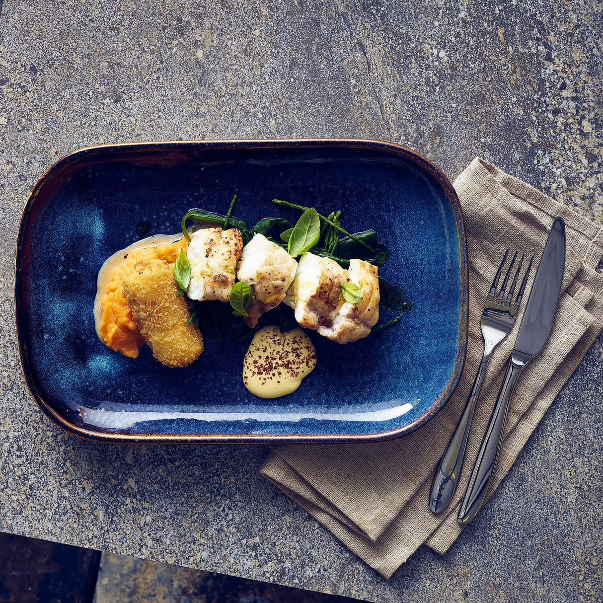 Blue plate with a dish of food on a gray surface with a napkin and cutlery.