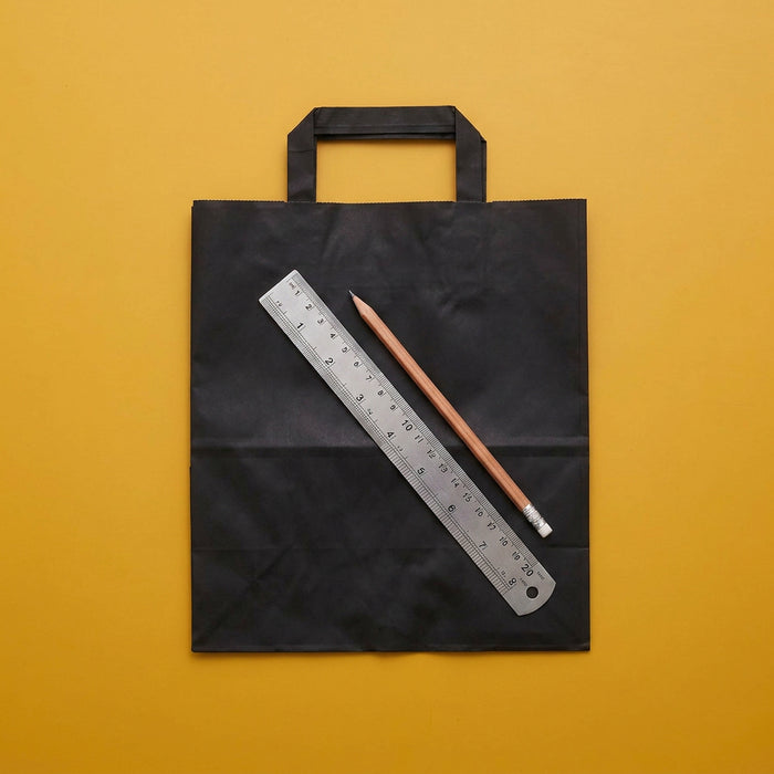 A black paper shopping bag with two handles, laid flat on a mustard yellow background, with a metal ruler and a wooden pencil placed diagonally across the front.
