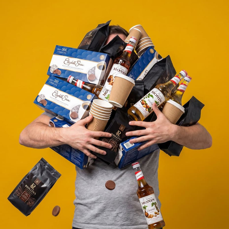 A man in a gray t-shirt is surrounded by a chaotic pile of coffee-themed products, including boxes of Épices Sucre, bottles of Monin syrup, paper cups, and coffee beans, set against a solid yellow background.