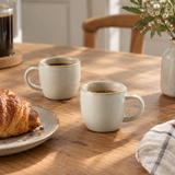Two speckled light beige ceramic coffee mugs filled with black coffee on a wooden table, styled with a croissant on a plate, a glass of dark coffee, and a vase of flowers.