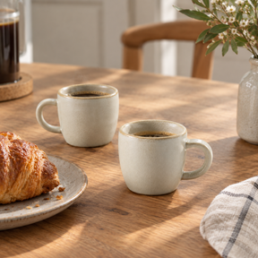 Two speckled light beige ceramic coffee mugs filled with black coffee on a wooden table, styled with a croissant on a plate, a glass of dark coffee, and a vase of flowers.