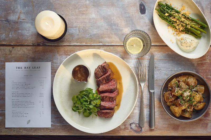 Overhead view of a plated steak dish on a rustic wooden table, with a menu card, candle, lemon wedge, asparagus, potato dish, and cutlery arranged around the main plate.