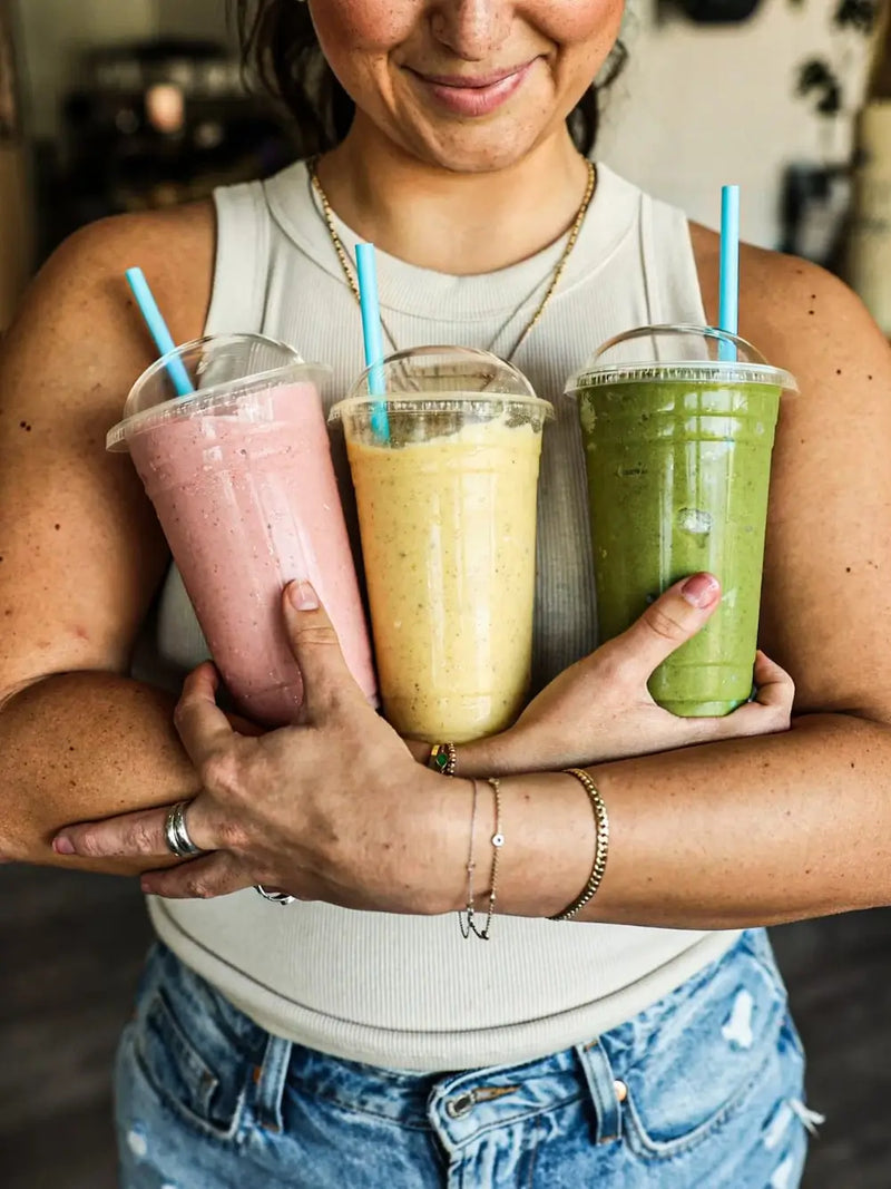 Person holding three colorful smoothies with straws in a casual setting