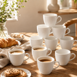A set of white ceramic coffee cups arranged on a wooden table in a warm, sunlit breakfast setting, with croissants, cookies, and a glass of coffee nearby.