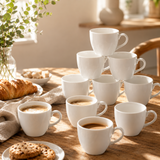 A set of white ceramic coffee cups arranged on a wooden table in a warm, sunlit breakfast setting, with croissants, cookies, and a glass of coffee nearby.