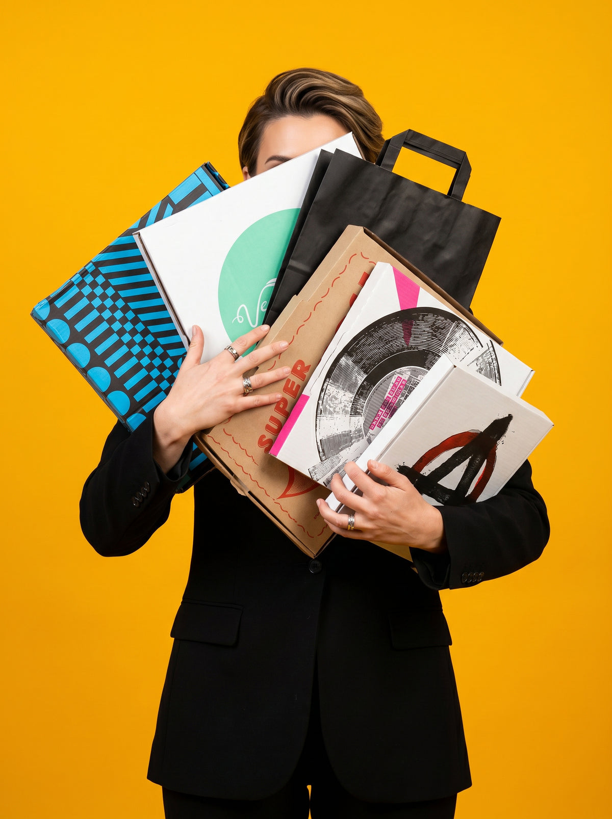A person in a black blazer holds a large stack of colorful vinyl record sleeves, a black shopping bag, and patterned folders against a solid yellow background.