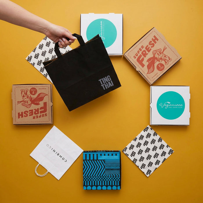 A hand holds a black paper shopping bag printed with the brand name 'TING THAI' in white, arranged on a bright yellow background with several branded food delivery boxes and paper bags.