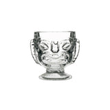 A clear glass with a Tiki-style head design, featuring a happy face on one side and a sad face on the reverse side, with a footed base.