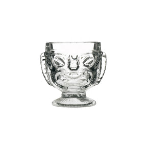 A clear glass with a Tiki-style head design, featuring a happy face on one side and a sad face on the reverse side, with a footed base.