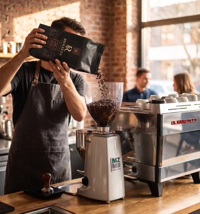 A barista in a dark apron pours coffee beans from a black MZ Coffee Roasters bag into a white and silver MZ Mini Pro grinder in a warm, rustic cafe setting.