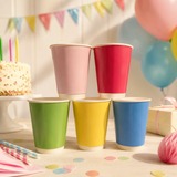 Six paper party cups in pink, red, green, yellow, and blue, stacked in a pyramid on a decorated birthday table with balloons, confetti, and a cake.