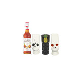 A Monin syrup bottle with orange liquid next to three decorative tumbler glasses: two white skull-shaped glasses and one black tiki-style glass with a pineapple design, on a plain white background.