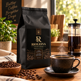A black bag of Rioléeva coffee beans stands on a wooden table in a warm, sunlit kitchen or cafe setting. A cup of black coffee on a saucer sits in front, with scattered coffee beans, a bowl, a plant, and a French press in the background.
