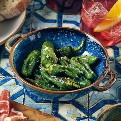 Blue ceramic bowl filled with green peppers on a decorative tablecloth.