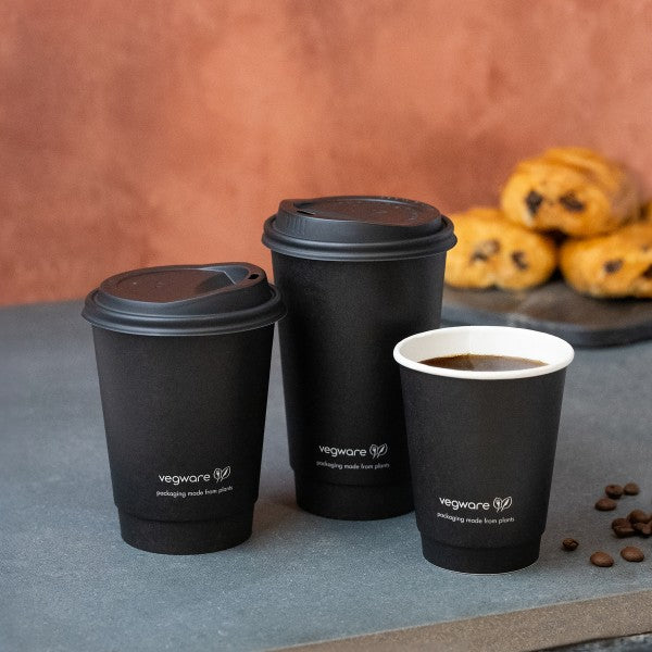 Black paper cups with lids on a gray surface, with 'vegware' branding, and pastries in the background.