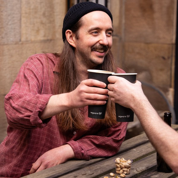 Man with long hair and beard holding two black coffee cups outdoors.