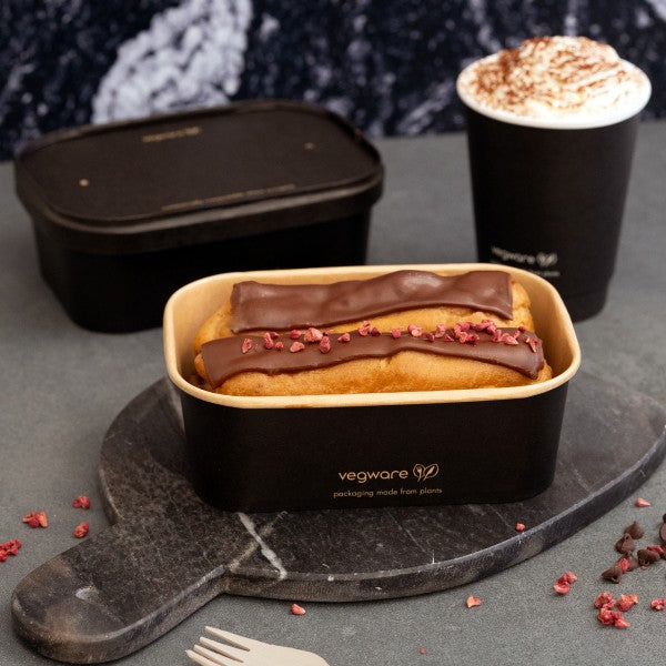 Eclairs in a black Vegware container on a dark surface with a cup of hot chocolate.