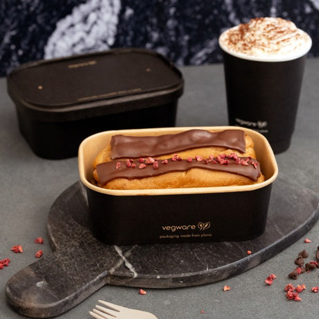 Eclairs in a black Vegware container on a dark surface with a cup of hot chocolate.